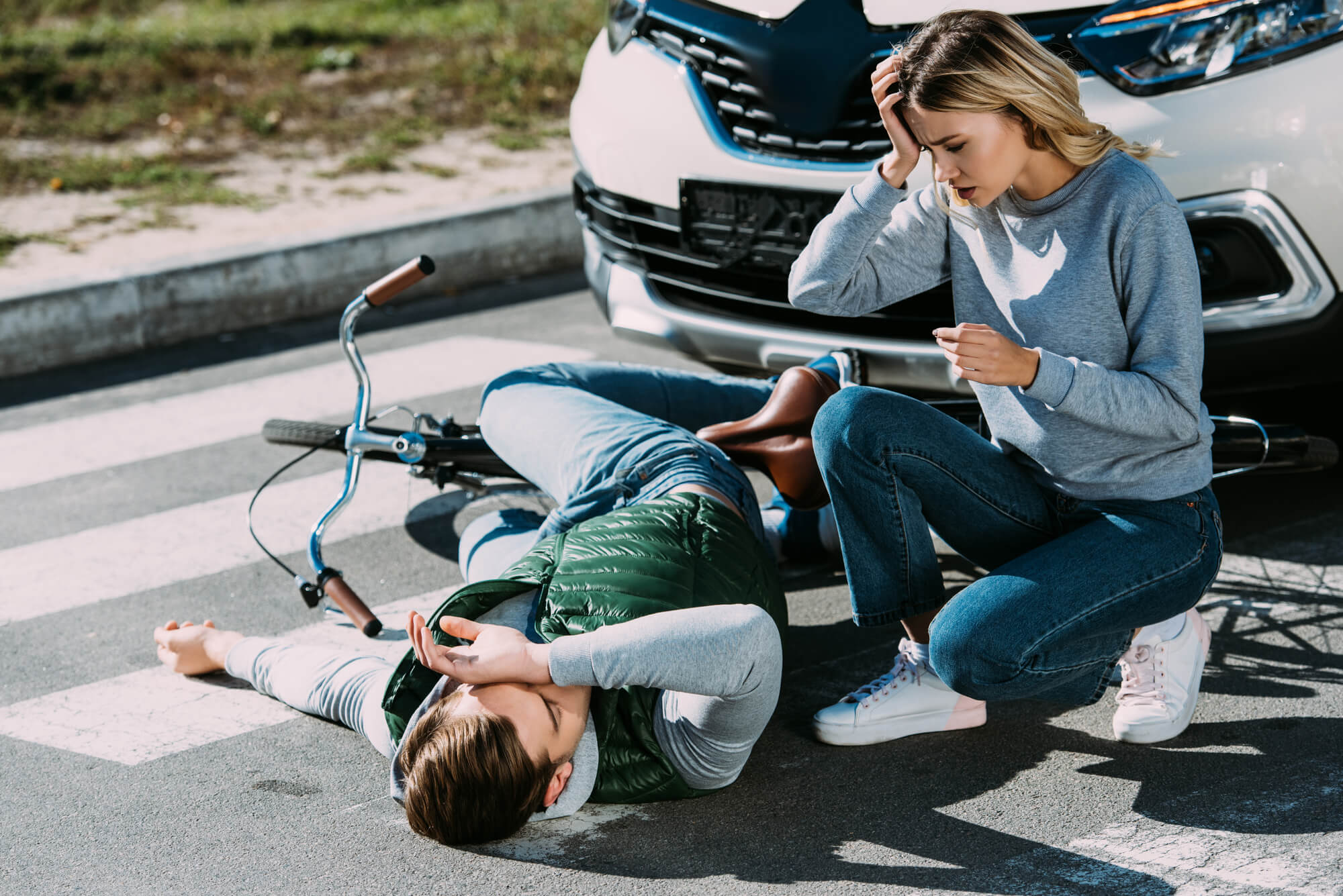 Shocked young woman looking at injured cyclist lying with bicycle at car accident